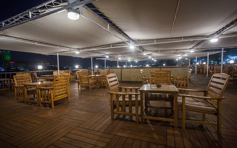 Open-air dining deck aboard the M/S City & Sea Adonis at night with wooden furniture and city lights.