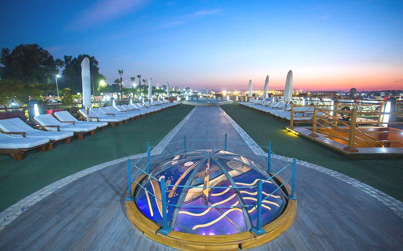 Sun deck with loungers, umbrellas and illuminated lounge area at dusk aboard cruise ship.