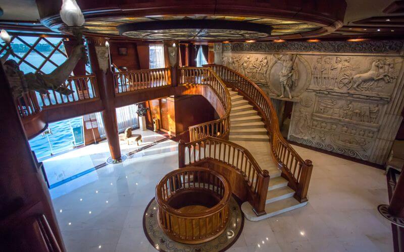Elegant multi-level atrium with wooden railings and Egyptian relief wall art aboard cruise ship.